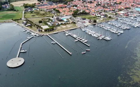 Luftaufnahme des Marstal Hafens mit vielen Segelbooten, die an Stegen liegen. Ein ovaler Steg mit Bänken ragt ins Wasser.