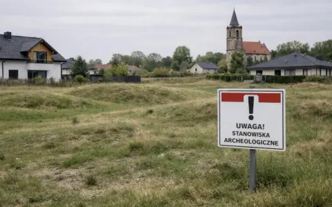 Tablica informuje o stanowiskach archeologicznych. W tle domy i kościół.