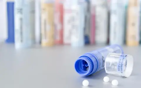Flacons de granules homéopathiques, certains en arrière-plan floutés. Un tube bleu ouvert laisse échapper quelques billes blanches. Une solution naturelle pour la dermite séborrhéique, comme une huile essentielle, pourrait être contenue dans ces remèdes.