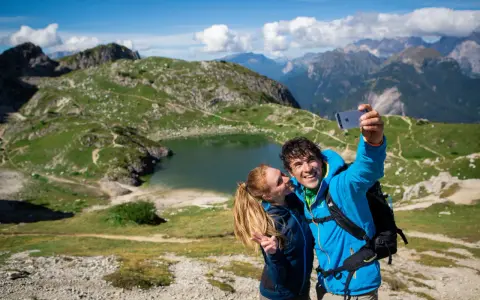Coppia felice si scatta un selfie con il lago Coldai sullo sfondo, circondato da montagne verdi e un cielo azzurro con nuvole bianche.