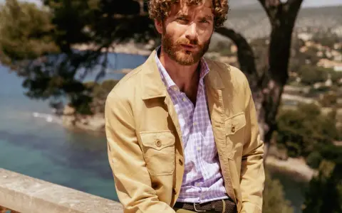 Un uomo con barba e capelli ricci indossa una camicia a quadri viola e una giacca color sabbia. Ottima idea su cosa mettere sopra la camicia uomo per un look casual chic.