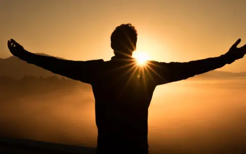 Silhouette eines Mannes mit ausgebreiteten Armen im Sonnenaufgang. Die Sonne bricht durch seine Silhouette und wirft Strahlen. Ein Symbol für Hoffnung und wie wird man Christ.