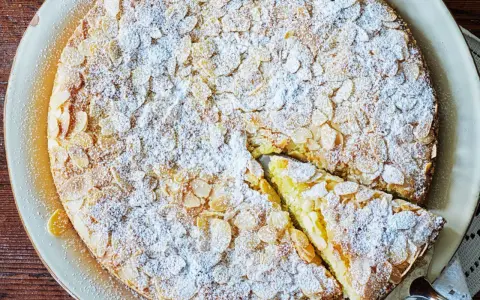 En saftig mandelkaka med citron, toppad med flagad mandel och pudrad florsocker. En bit är uppskuren.
