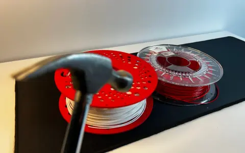 Młotek zbliża się do szpul z filamentem, testując wytrzymałość materiałów. Biały i czerwony filament czekają na próbę.