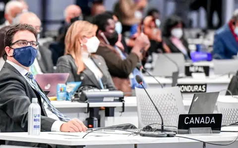 Teilnehmer der Klimakonferenz, darunter ein Mann mit Brille und Maske, sitzen an Tischen mit Laptops und Namensschildern wie "CANADA".