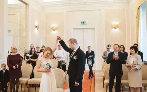 Para młoda świętuje swój ślub cywilny, trzymając się za ręce. W tle widać gości i rodzinę.
