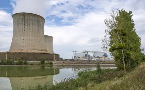 Kühltürme eines Atomkraftwerks in Frankreich, die Dampf abgeben. Ein ruhiger See spiegelt die Anlage wider, während am Ufer Bäume und Gras wachsen.