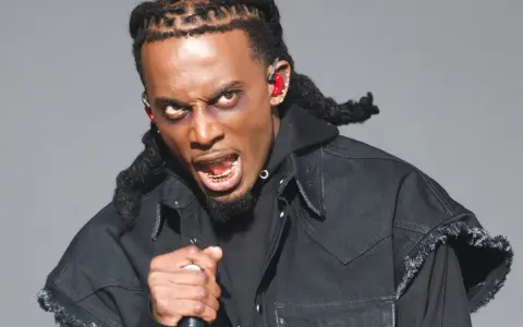 Playboi Carti, wiek nieznany, w czarnej kurtce z frędzlami, z mikrofonem w dłoni, z wyrazem twarzy pełnym pasji.