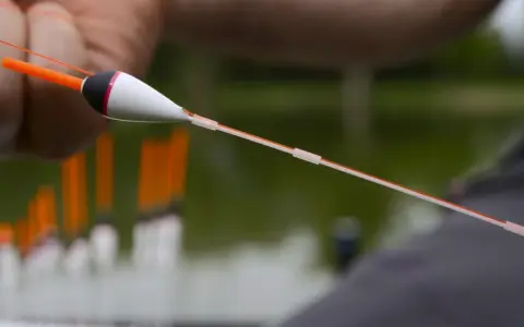 Zbliżenie na rękę trzymającą spławik wędkarski z pomarańczową antenką i biało-czarnym korpusem.