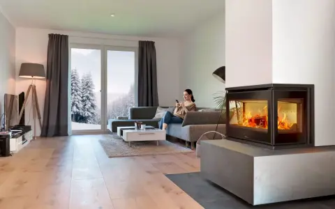 Mujer en sofá mira móvil junto a chimenea de cassette pellets con fuego vivo. Ventana muestra paisaje nevado.