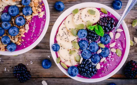 Zwei Schalen mit gesunden Bowls, gefüllt mit lila Smoothie, garniert mit Blaubeeren, Bananenscheiben, Brombeeren, Kürbiskernen und Granola.