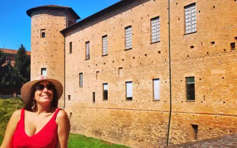 Donna sorridente con cappello di paglia davanti a un castello di mattoni rossi. Un'idea per cosa vedere a Piacenza in un giorno.