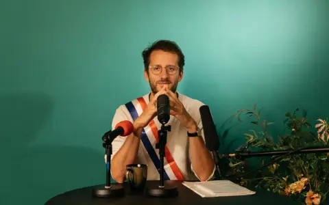 Un homme avec une écharpe tricolore, prêt à parler. Il est entouré de micros, peut-être pour un discours plein de langue de bois.