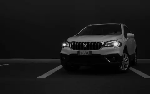 Białe Suzuki SX4 na ciemnym tle, z włączonymi światłami.