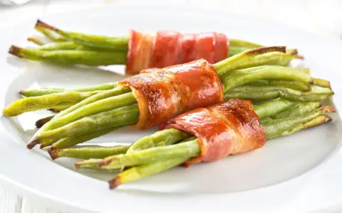 Drei Portionen Bohnen im Speckmantel auf einem weißen Teller. Der Speck ist knusprig gebraten und umhüllt die grünen Bohnen.