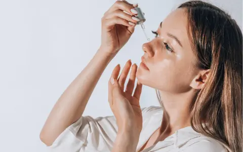 Frauenhand trägt Tropfen eines Serums auf ihr Gesicht auf. Ob Squalan oder Hyaluron, die Haut strahlt Feuchtigkeit aus.