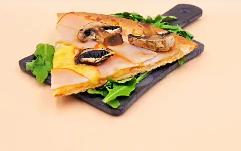 Kawałek pysznej pizzy capriciosa z szynką, serem i pieczarkami na ciemnej desce, ozdobiony rukolą.