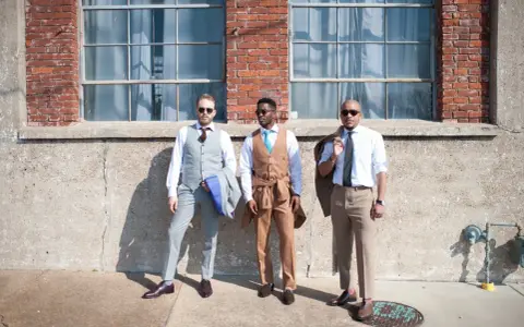 Three men in stylish suits embody cocktail chic attire. One wears a grey pinstripe suit, another a tan suit with a turquoise tie, and the third a light blue shirt with tan trousers.