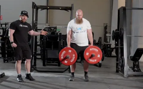 En man med skägg utför farmers walk med tunga vikter. En tränare visar hur övningen ska göras.