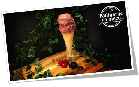 Dwa kule czekoladowych lodów białkowych w wafelku, ozdobione wiśniami i kawałkami gorzkiej czekolady.