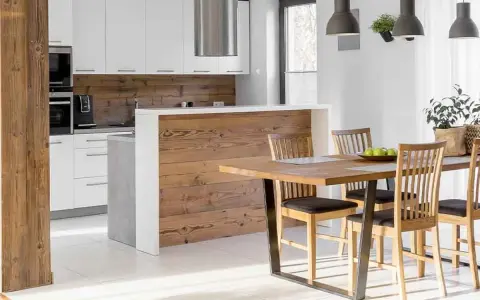 Cocina semiabierta al salón con barra de madera, encimeras blancas y electrodomésticos integrados. Mesa de comedor de madera con sillas.