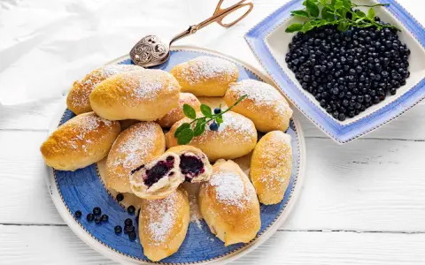 Pyszne jagodzianki posypane cukrem pudrem, obok miseczka pełna świeżych jagód. Idealne na letnie popołudnie.