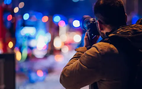 Mężczyzna z aparatem w ręku, uchwycony w trakcie nocnej fotografii. Rozmyte, kolorowe światła miasta tworzą magiczne tło.