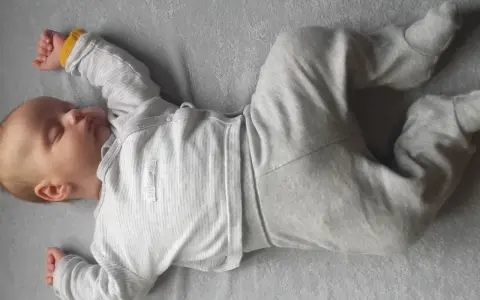 Śpiący noworodek w szarym pajacyku. Jego rączki i nóżki są ułożone w lekkiej asymetrii, co jest naturalne dla niemowląt.