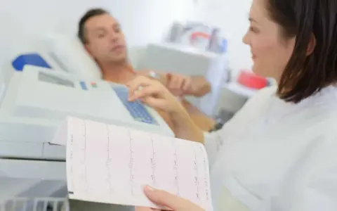 Pielęgniarka analizuje zapis EKG, który pokazuje, jakie są badania serca. Pacjent leży podłączony do aparatury.