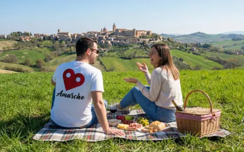 Coppia in un prato verde con vista su un borgo collinare. Perfetto per scoprire cosa fare a Pasquetta nelle Marche con un picnic.