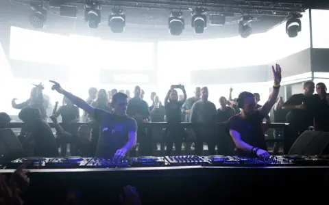 Dos DJs actuando en un club con público al fondo.
