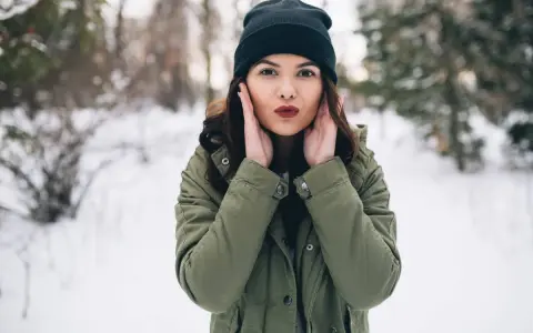 Kobieta w zielonej kurtce i czarnej czapce zimowej, na tle zaśnieżonego lasu.
