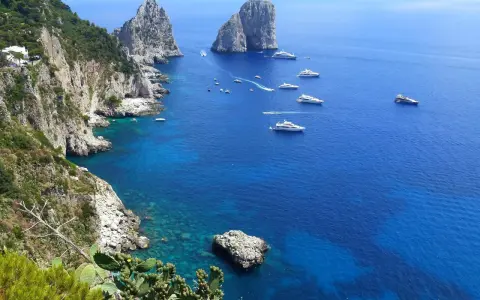 Scogliere verdi e rocce imponenti si affacciano sul mare blu intenso, con yacht che navigano. Un'immagine che ispira su come raggiungere Capri da Napoli.