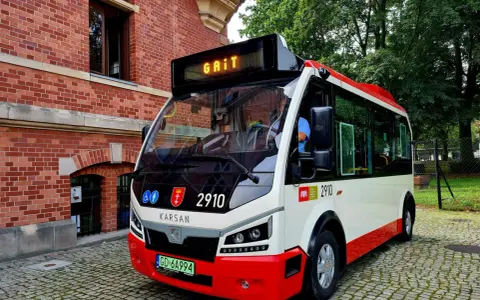 Nowy autobus 100 z napisem "GAIT" na wyświetlaczu, zaparkowany na brukowanej ulicy obok ceglanego budynku.