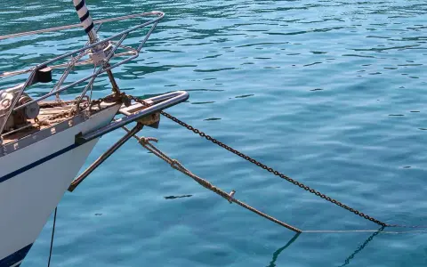 L'étrave d'un voilier blanc est ancrée dans une eau bleue et claire. La chaîne et la corde sont bien visibles, prêtes à mouiller beaucoup.