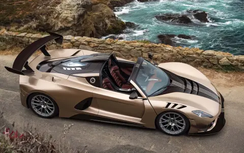 Der Hennessey Venom F5 Roadster in Goldbraun steht an einer felsigen Küste. Sein aggressives Design mit großem Heckspoiler und dunklen Streifen zieht alle Blicke auf sich.