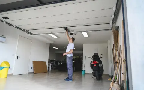Un homme ajuste le moteur de la porte de garage électrique. Un scooter et des outils sont garés à proximité.