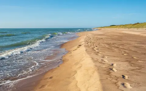 Spokojne, dzikie plaże nad Bałtykiem. Fale delikatnie obmywają piaszczysty brzeg, a na piasku widoczne są ślady stóp.
