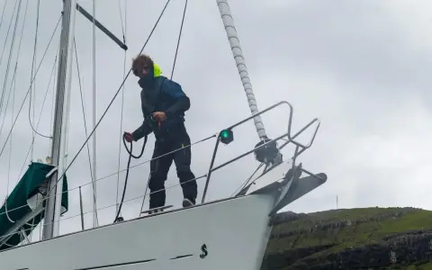 Un marinier, les amarres avis, manœuvre sur le pont d'un voilier blanc, face à une côte rocheuse verdoyante.