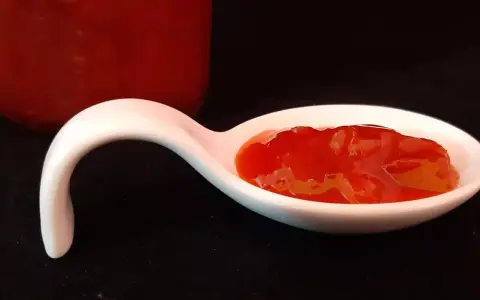 Une cuillère blanche présente une portion de confiture fraise rhubarbe, d'un rouge vif et appétissant. Un pot de confiture est visible en arrière-plan.