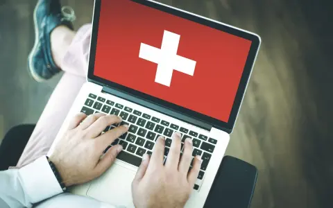 Osoba pisząca na laptopie z flagą Szwajcarii na ekranie.