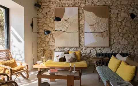 Salon avec mur en pierre, deux tapisseries texturées, fauteuil en rotin, banquette jaune et coussins aux motifs ethniques. Une ambiance deco campagne boheme chaleureuse.