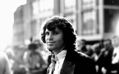 Jim Morrison, mit seinem charakteristischen lockigen Haar und einem nachdenklichen Blick, steht im Gegenlicht.