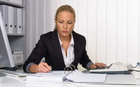 Kobieta w biurze pracuje nad dokumentami, używając kalkulatora i długopisu. Na biurku leżą stosy papierów i segregatorów, a w tle widać regał z dokumentacją. Może przygotowuje weksle.