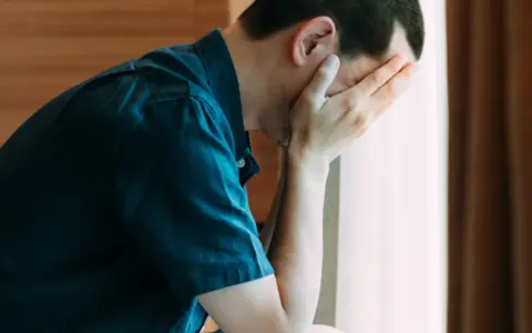 Mężczyzna w ciemnej koszuli ukrywa twarz w dłoniach, pogrążony w smutku. Może to być reakcja na ból, jak zaczerwienione jądra, lub inne cierpienie.