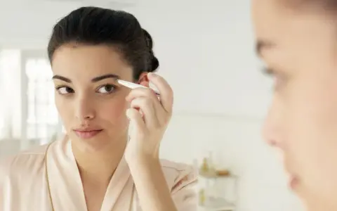 Une jeune femme ajuste ses sourcils broussailleux avec une pince à épiler devant un miroir, dans une tenue de chambre satinée.