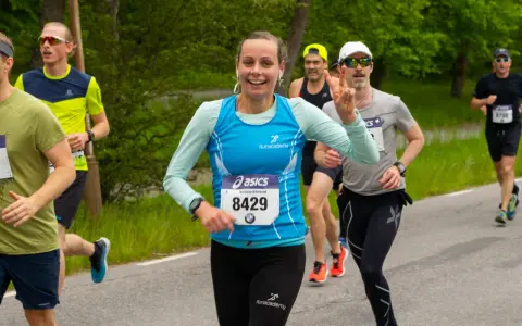 Glada löpare i ett 10 km lopp. En kvinna med nummer 8429 ler och gör segertecken.