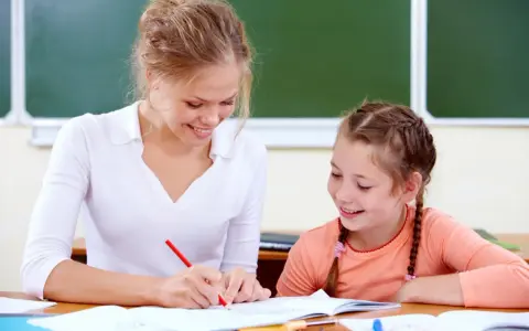 Uśmiechnięta nauczycielka wspomagająca z kwalifikacjami pomaga uczennicy w lekcjach przy biurku.