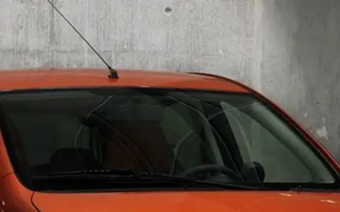 Pomarańczowy samochód z przyciemnianymi szybami z przodu, z antenką na dachu, stoi przy betonowej ścianie.