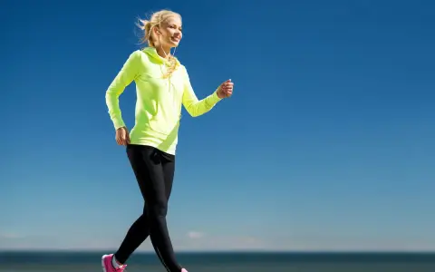 Uśmiechnięta blondynka w neonowej bluzie i czarnych legginsach uprawia power walking nad morzem.